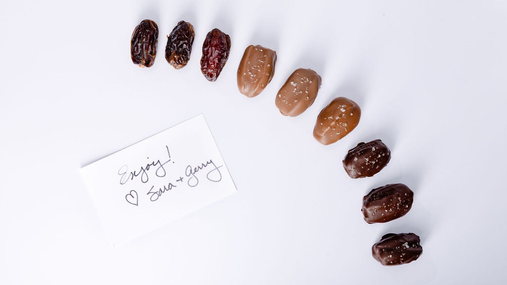 An arc of plain, milk chocolate, and dark chocolate–covered dates is arranged beside a handwritten note that says, “Enjoy! ♥ Sara + Gerry.” on a white background.
