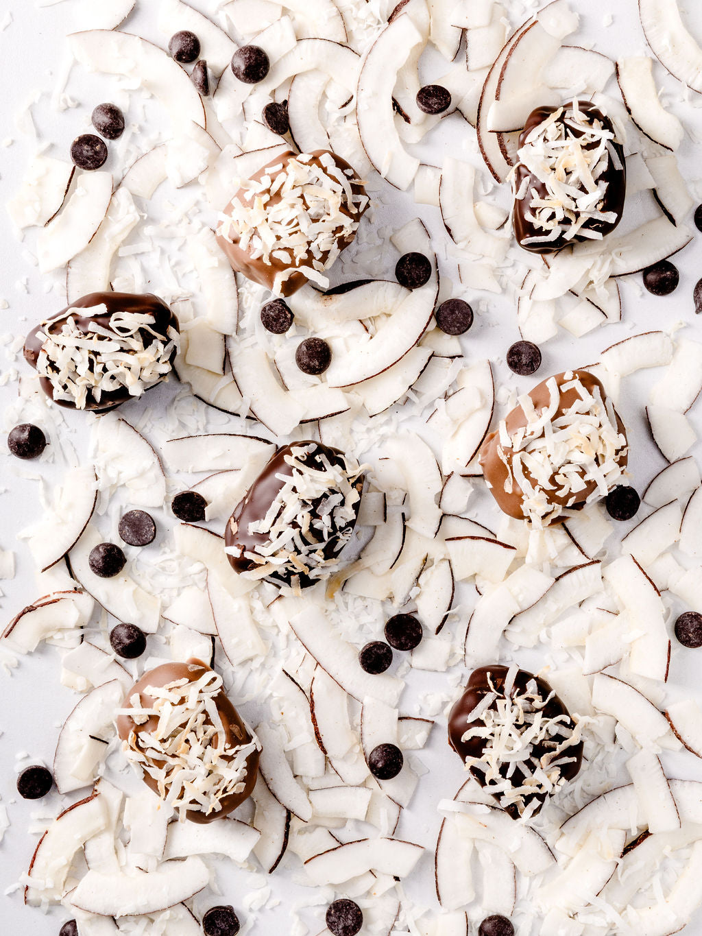 Chocolate-covered medjool dates topped with shredded coconut, surrounded by coconut pieces and shavings, and chocolate chips on a white surface.