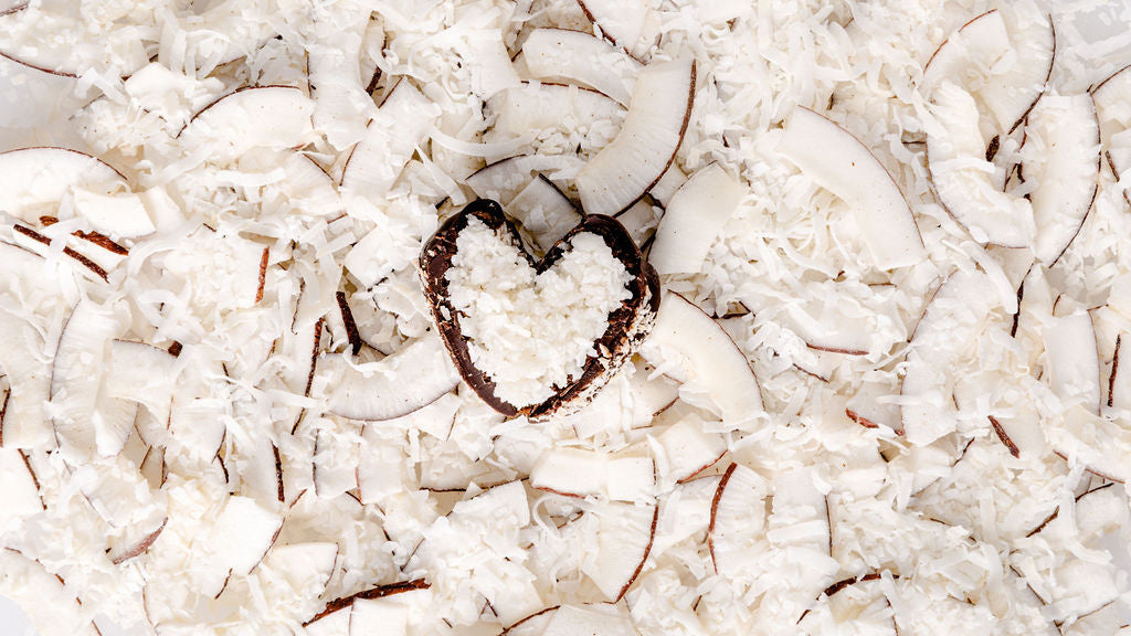A heart-shaped chocolate shaped medjool date filled with shredded coconut sits on a bed of coconut flakes and slices.
