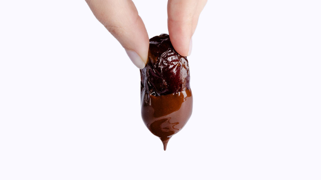A hand holds a date partially dipped in melted chocolate, with a drip forming at the bottom on a white background.