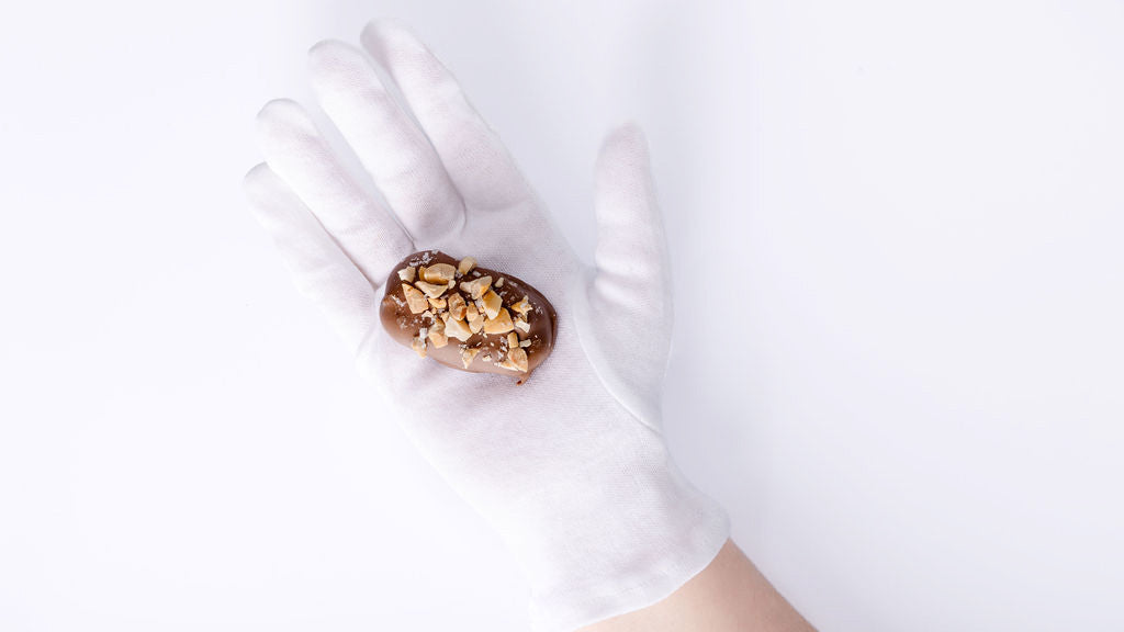 A white gloved hand holds a chocolate-covered medjool date with chopped peanuts on a white background.