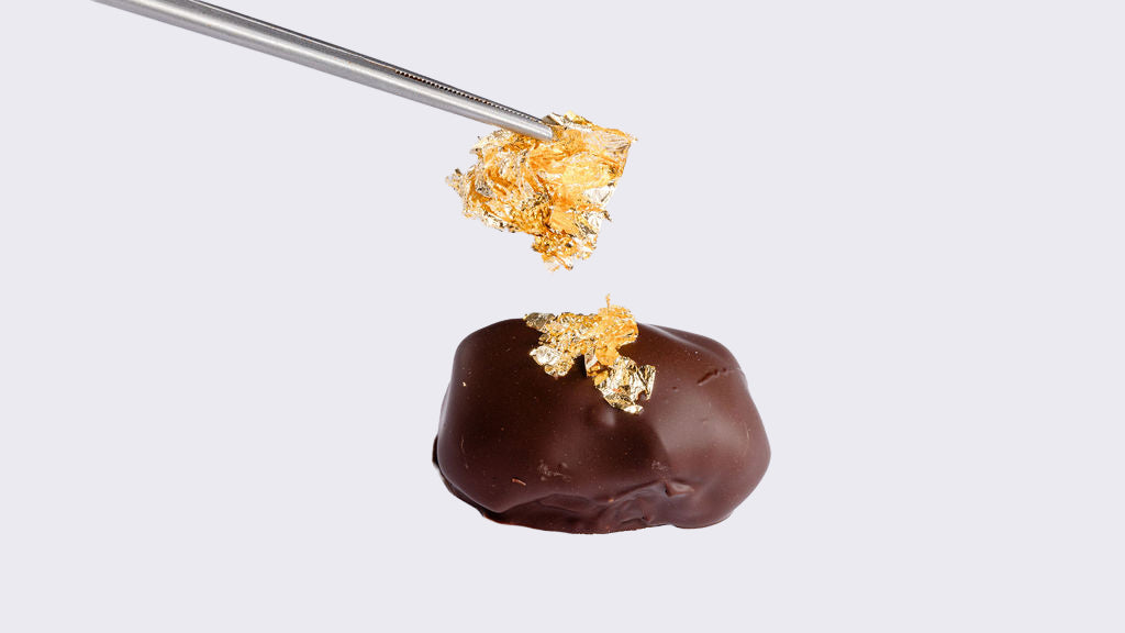 A piece of gold foil held by tweezers is being placed on top of a chocolate-covered medjool date on a white background. 